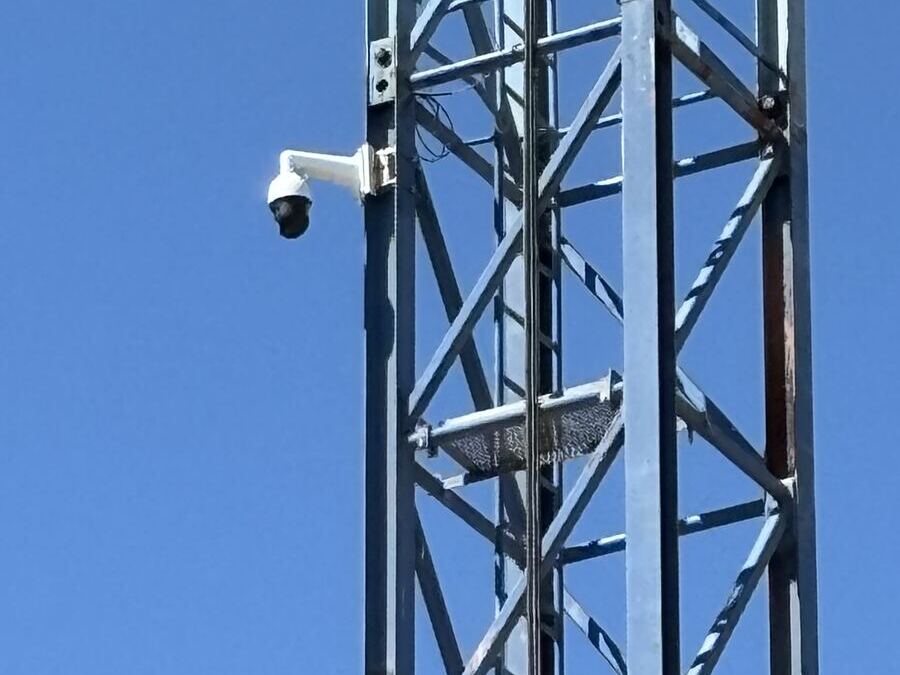 Camera mounted high on a crane lattice against a clear sky